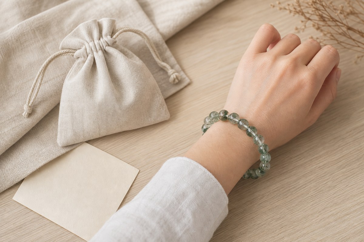 Green phantom quartz bracelet held near a neutral gift pouch on ivory linen and wood