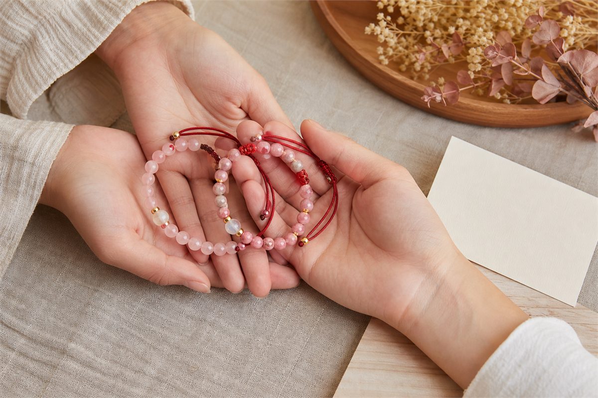Paired red string and rose quartz bracelets held by two hands in soft warm light