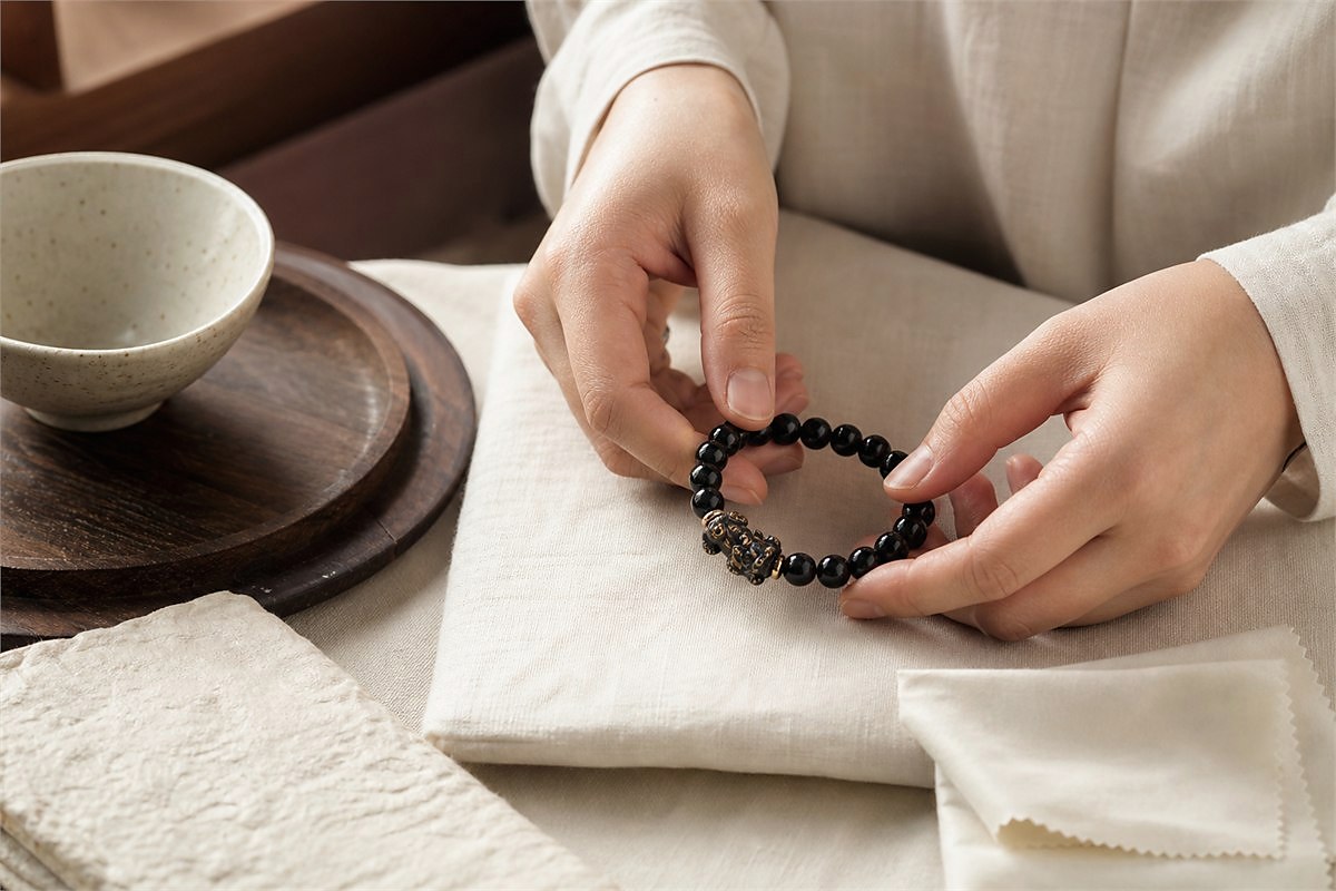 Hands preparing a black Pixiu bracelet on clean ivory linen beside a ceramic bowl and soft cloth