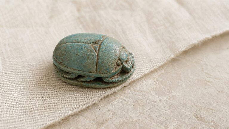 Blue-green ancient Egyptian faience scarab amulet on warm ivory linen and pale limestone.