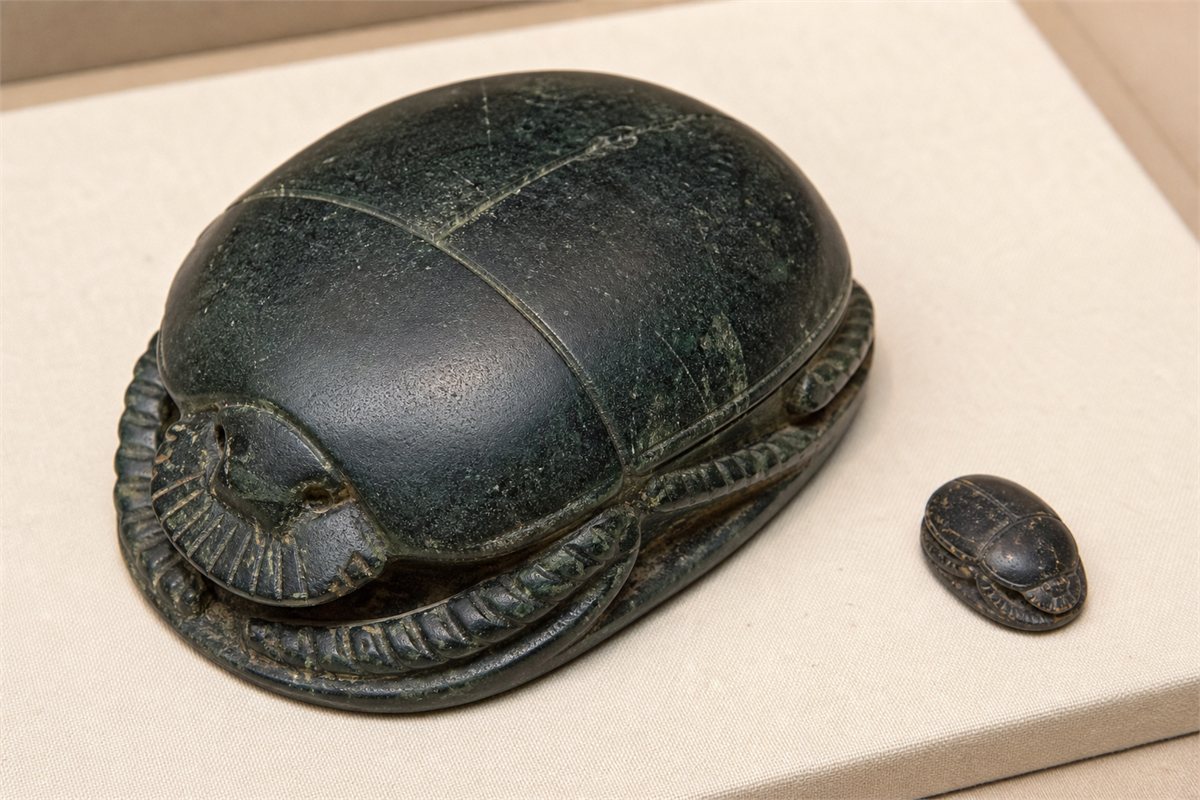 Large dark green stone heart scarab beside a much smaller ordinary scarab on a museum mount