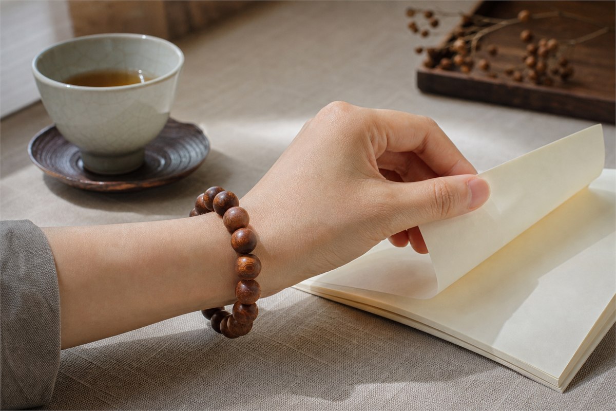Adult wrist wearing a brown wood bead bracelet while turning a blank cream paper page beside tea