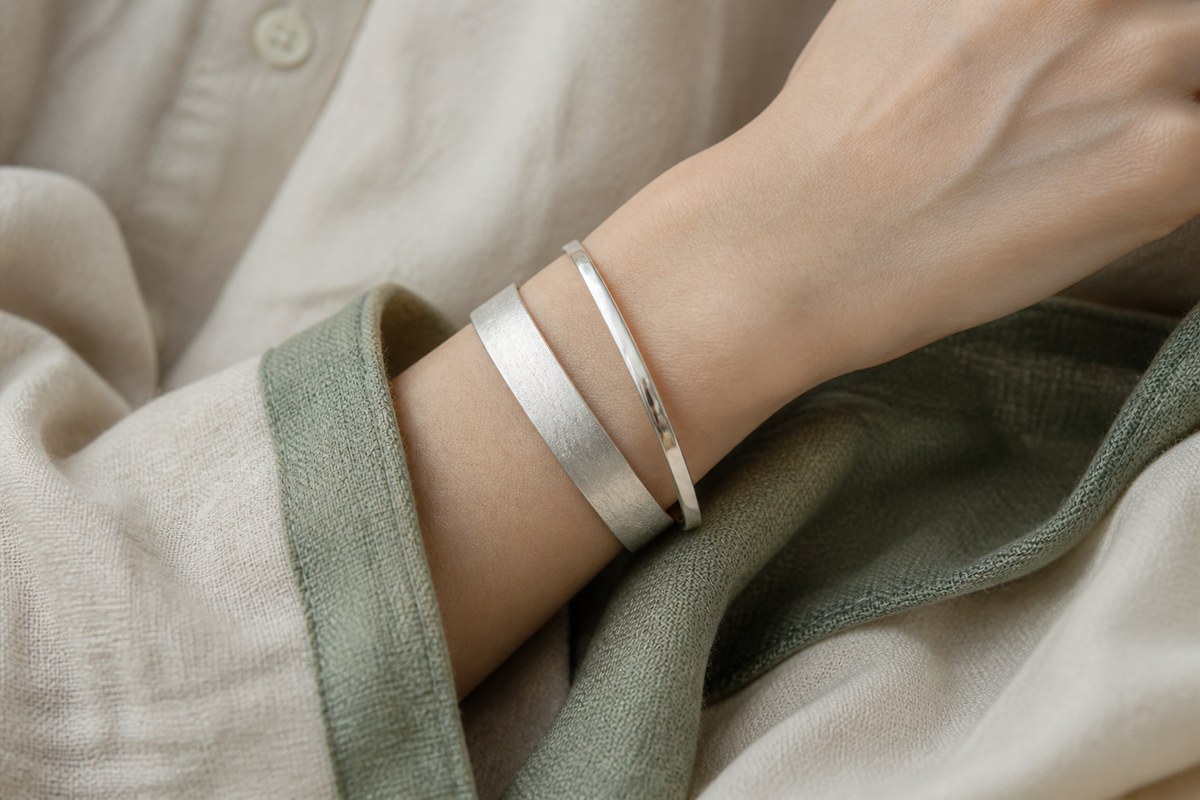 A wrist wearing layered silver bangles with a muted jade green textile and pale linen sleeve.