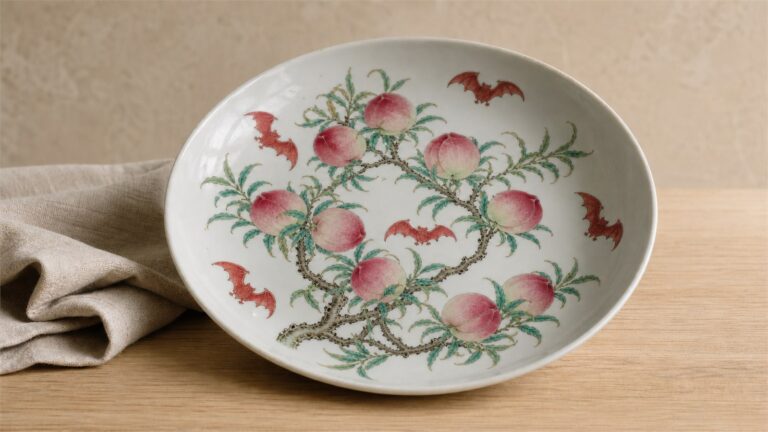 White porcelain dish painted with nine peaches and five red bats on a warm neutral tabletop