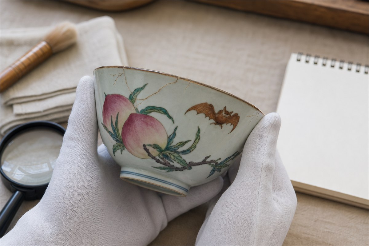 Gloved hands inspecting a peach-and-bat porcelain dish beside a loupe and blank notebook