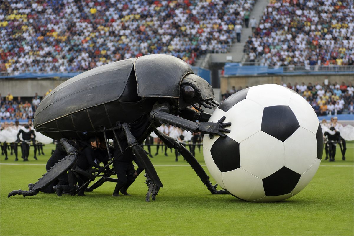 Giant constructed dung beetle stage puppet pushing an oversized football in a stadium.