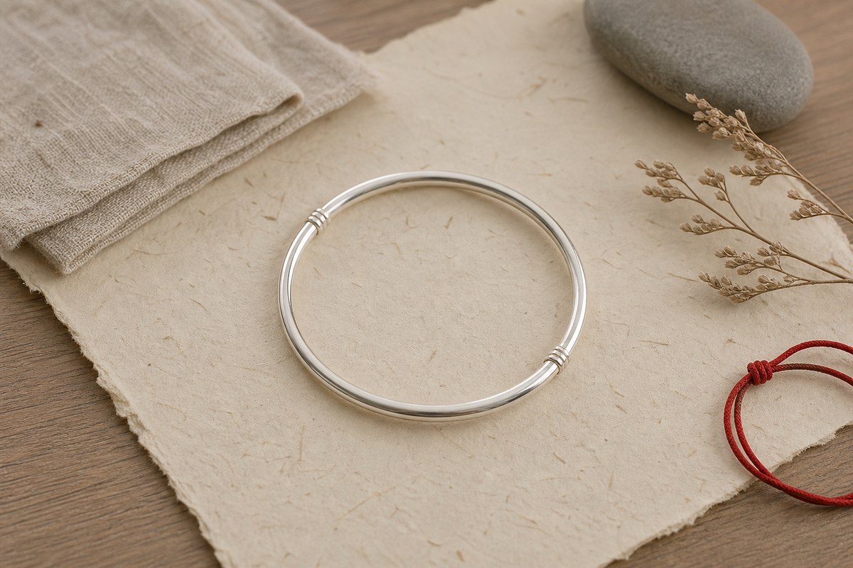 A simple polished silver bangle arranged on handmade paper with linen, stone, and a small red cord accent.