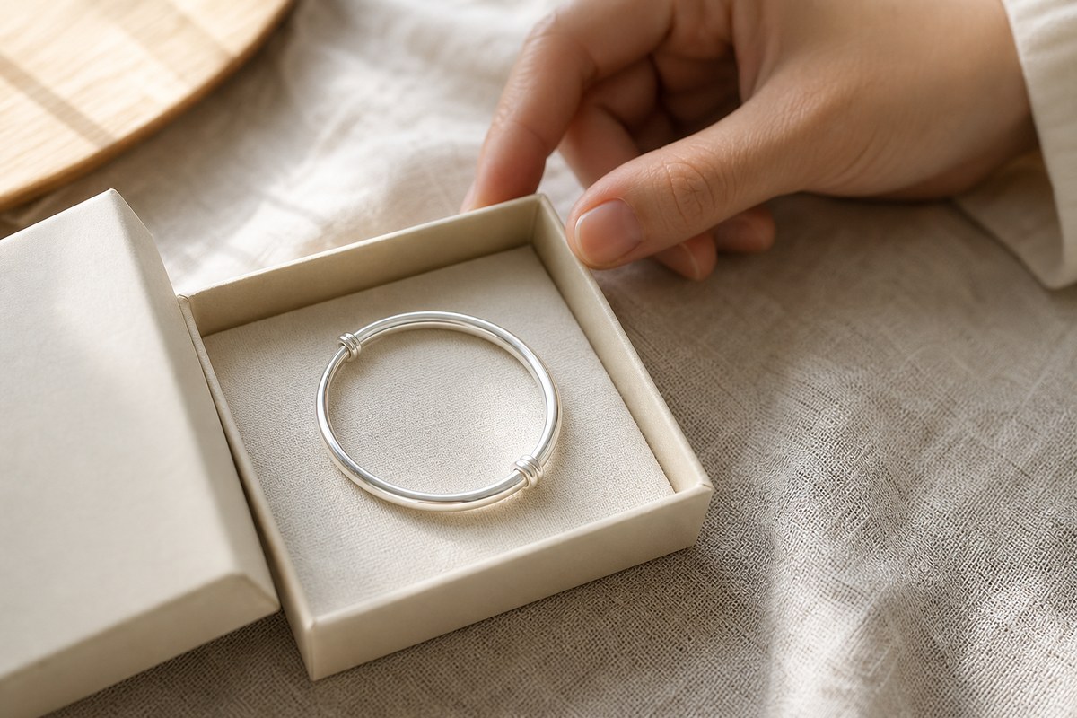 A small silver bangle resting in a cream gift box beside a hand in soft natural light.