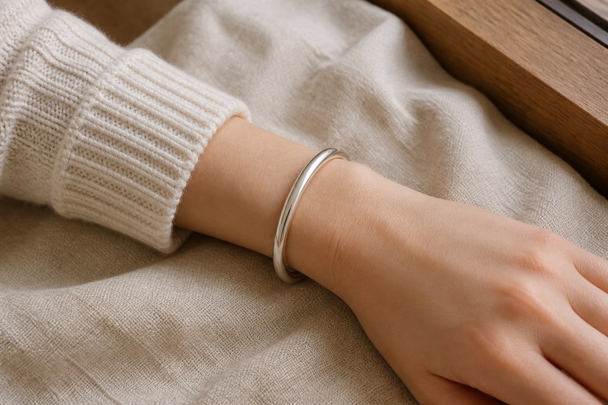 A simple silver bangle worn on a wrist with a cream knit sleeve and neutral linen background.