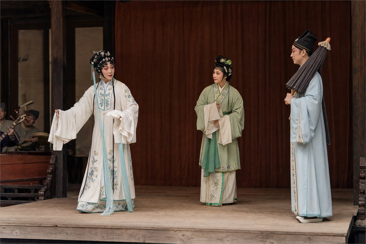 Three anonymous opera performers in white, green, and scholar costumes rehearsing with an umbrella on a bare wooden stage