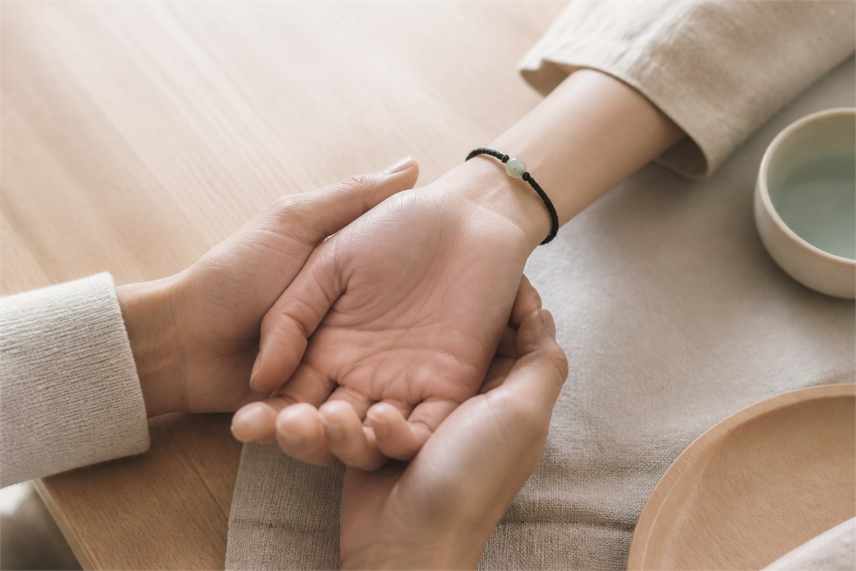 One person gently holding another hand wearing a slim black cord bracelet with a pale green bead