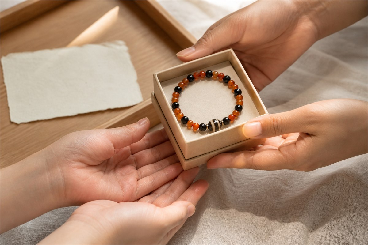 Hands offering a gift box containing a bracelet of orange-red carnelian, black chalcedony, and one banded onyx accent bead