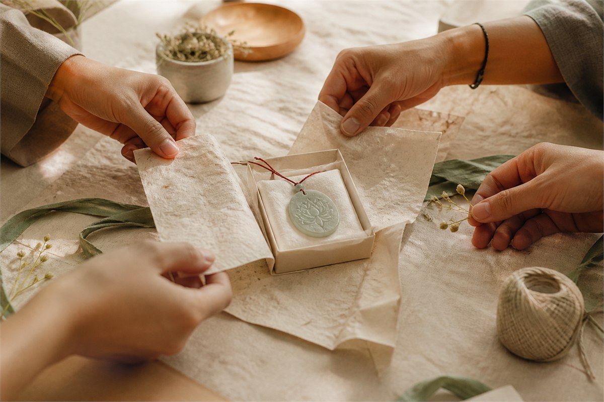 Four people wrapping a muted jade lotus pendant together on warm handmade paper
