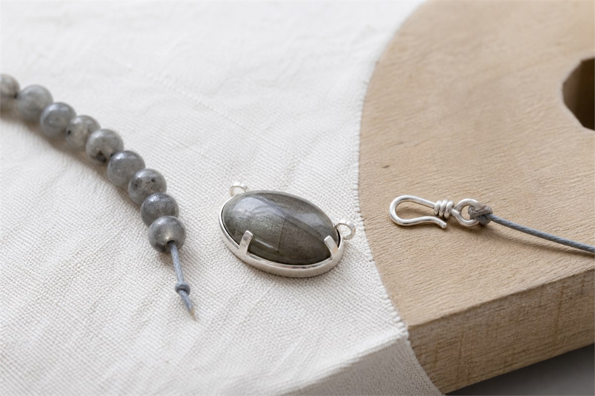 Gray feldspar cabochon in a silver-tone protective setting beside drilled beads, cord, and a small clasp