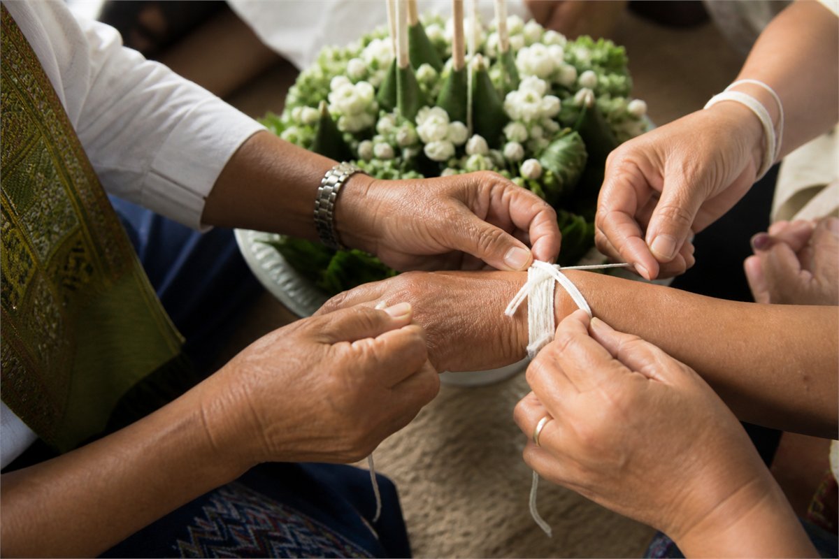 Several community members tie white cotton threads around an adult’s wrist beside a floral banana-leaf centerpiece.