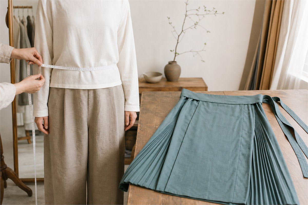 Tailor measuring the intended waistband position beside a teal pleated ma mian qun on a worktable