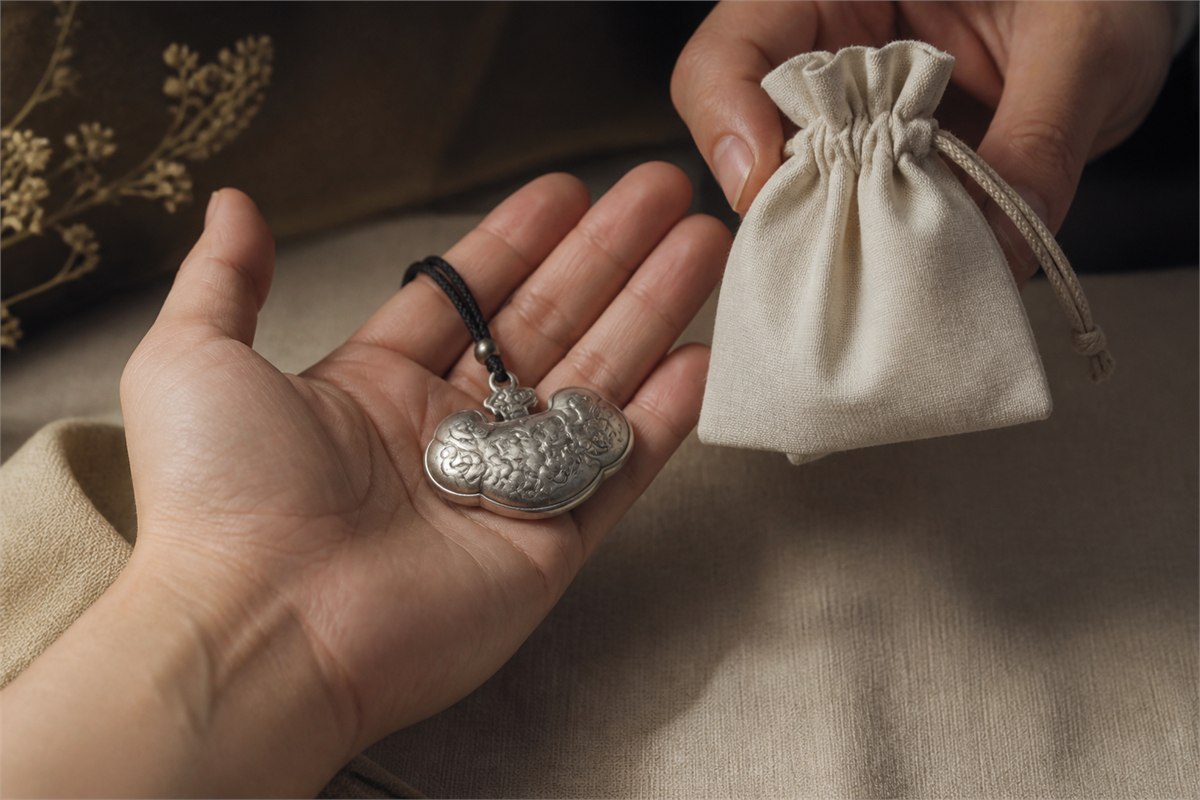 Silver ruyi-cloud pendant resting in an open palm while another hand offers a plain ivory fabric pouch