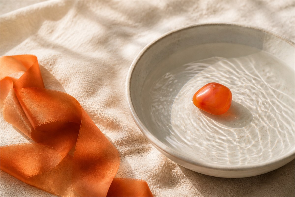 Orange carnelian stone in a shallow bowl of rippling water beside an orange ribbon on cream linen