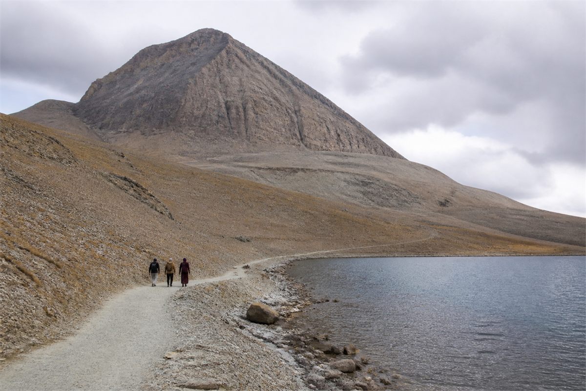Three distant walkers following an established path beside a high-altitude lake and mountain