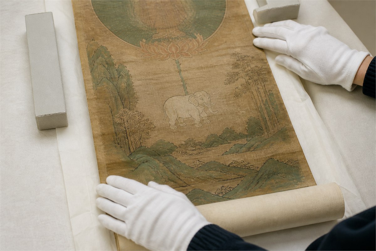 Gloved conservator hands examining an aged East Asian scroll with an elephant beneath a lotus canopy