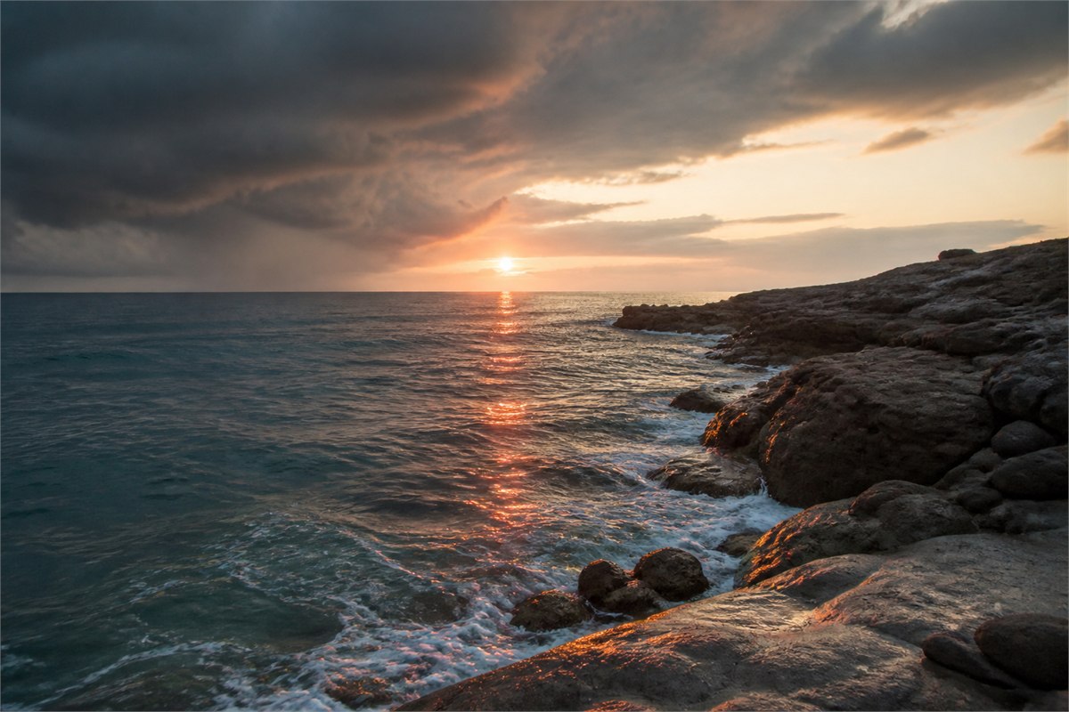 Sunset reflection crossing sea water beside dark coastal rock under opening clouds