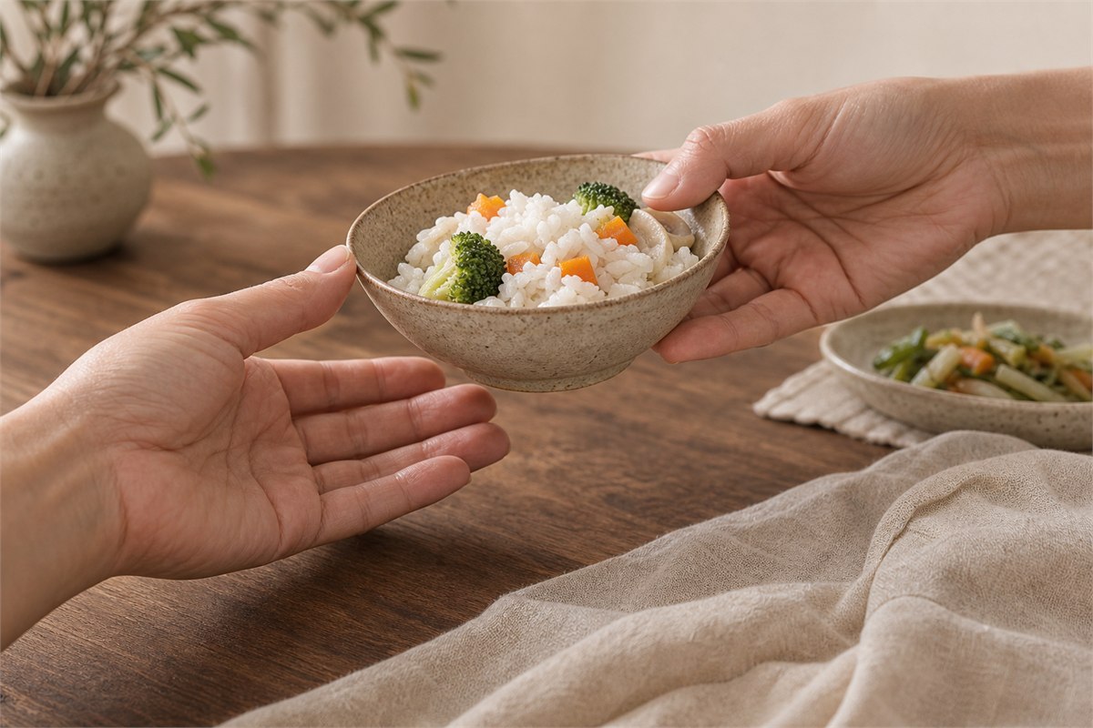Two hands passing a ceramic bowl of rice and vegetables across a warm wooden table.