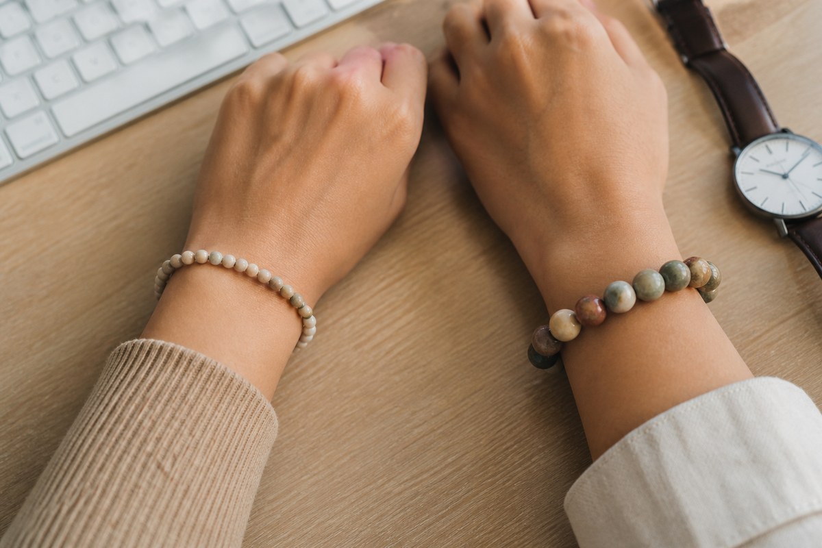 Two adult wrists wearing separate small pale beads and larger muted stone beads beside a keyboard and plain watch