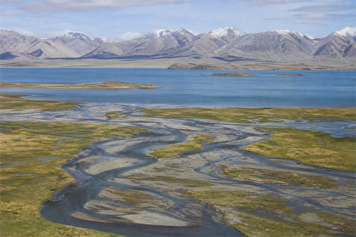 Braided river channels crossing a wetland toward a blue high-altitude lake and mountain range