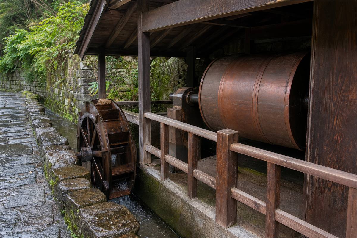 Wooden water wheel connected to a sheltered copper prayer wheel beside a shallow channel