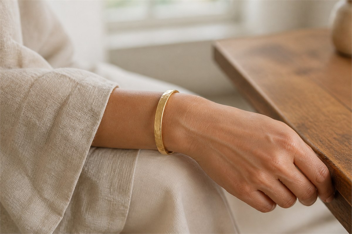 Neutral adult wrist wearing one subtly hammered matte gold bangle with linen