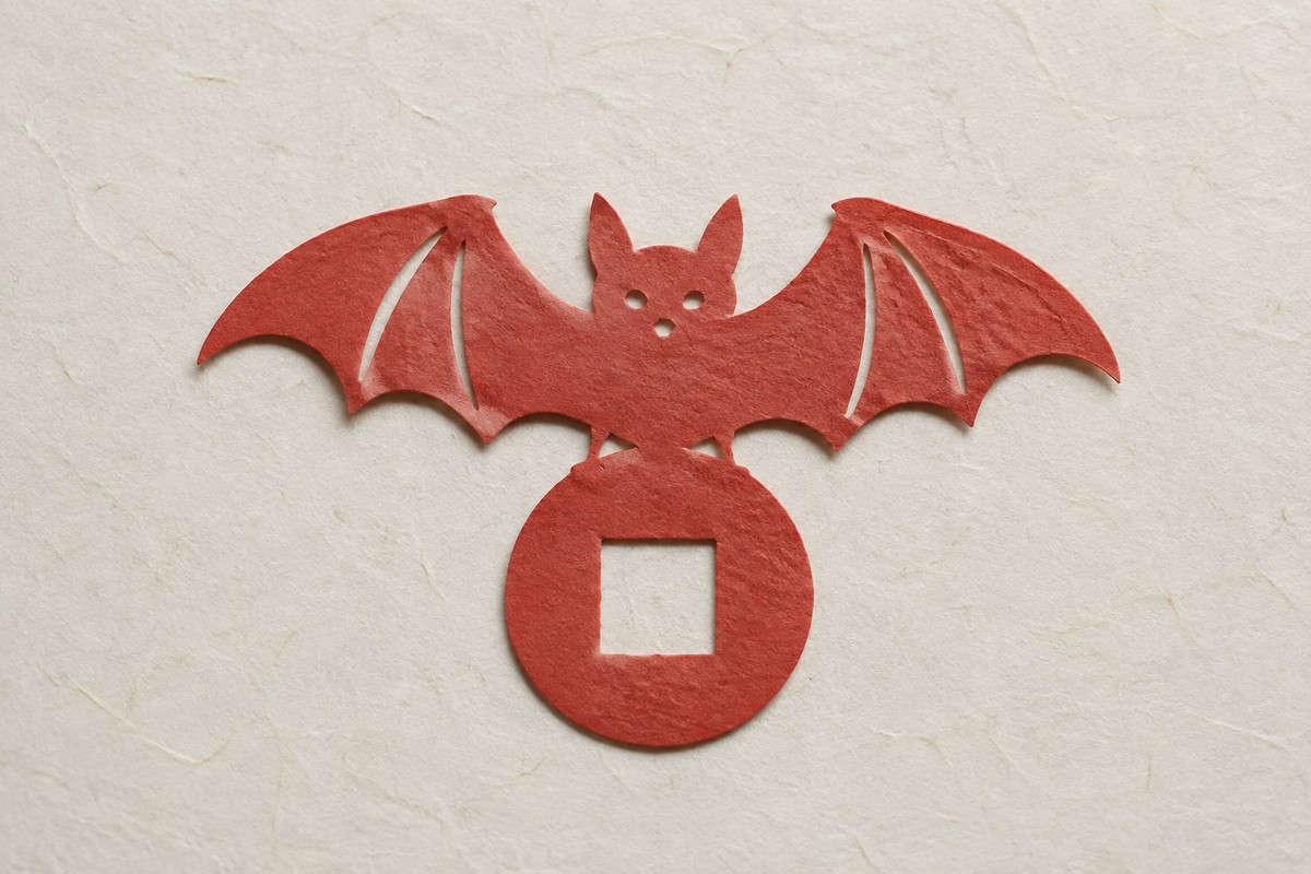 Muted red paper cut of a bat and a square-holed coin on pale handmade paper.