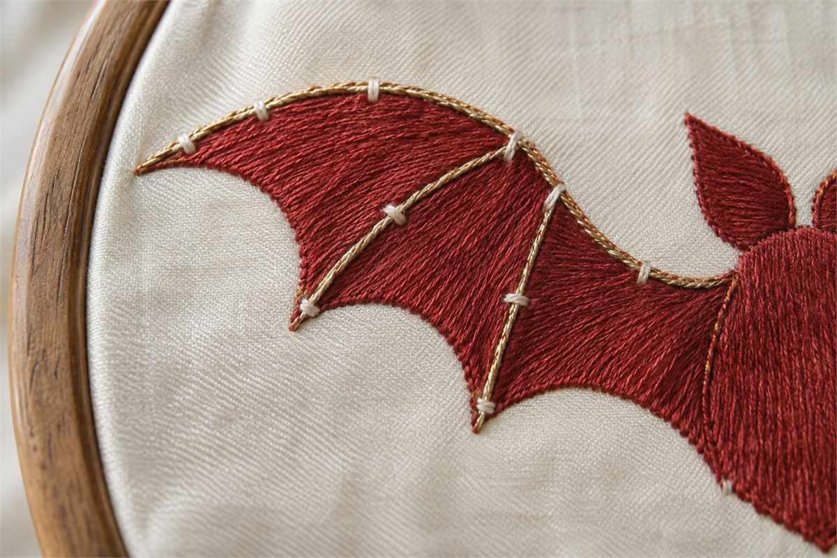 Gold-colored thread secured by small stitches around a red embroidered bat wing.