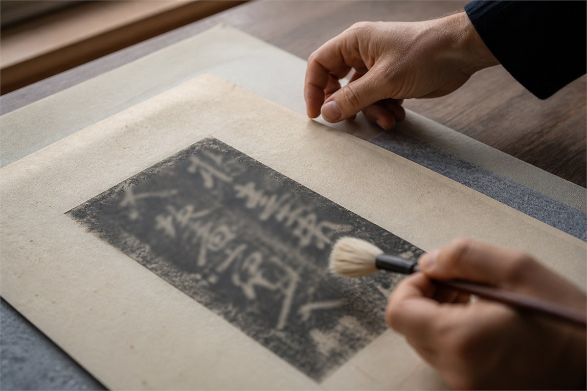Hands examine an intentionally blurred black-ink calligraphy rubbing with a soft brush.