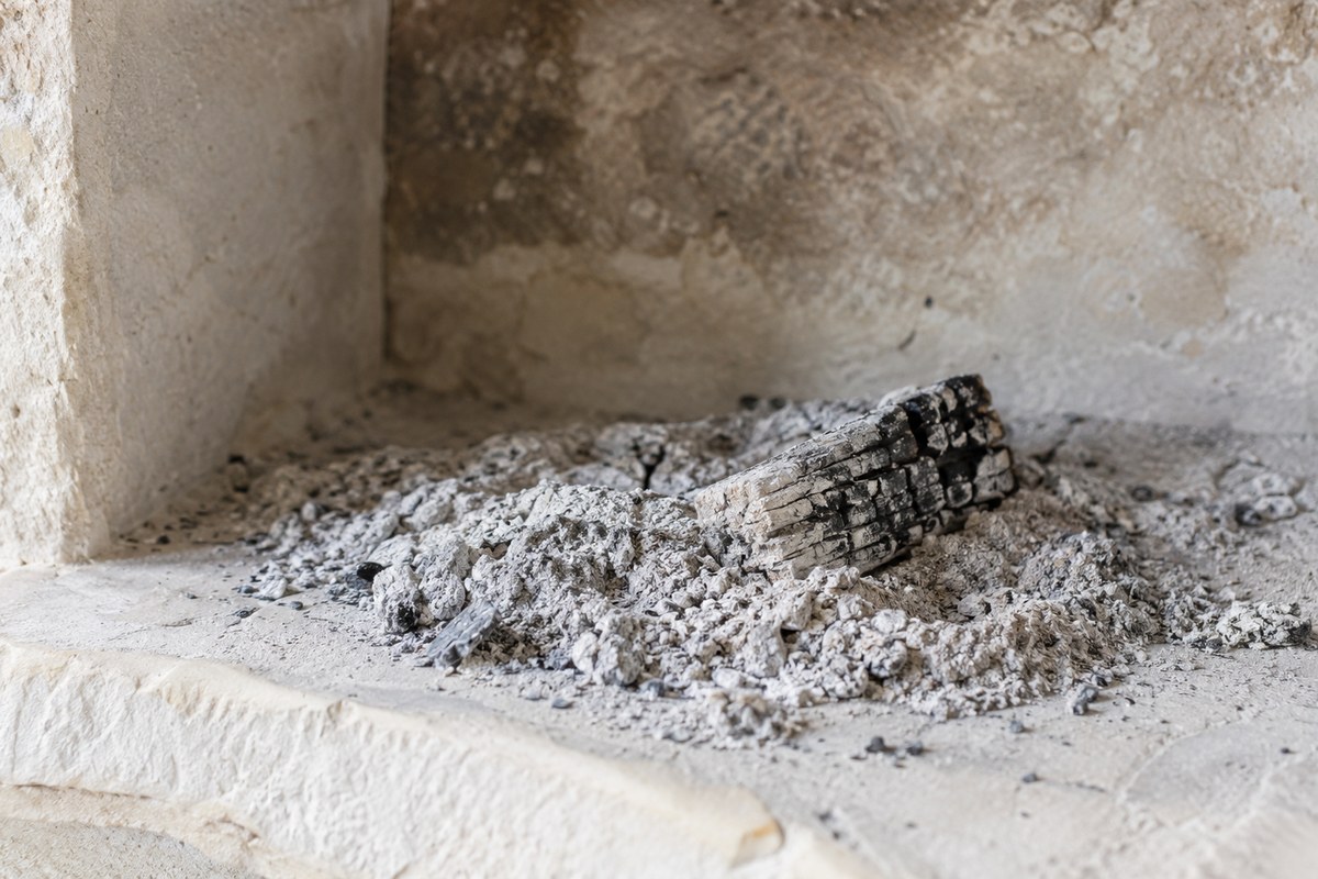 Cool pale gray wood ash and a small charred wood remnant lie in a shallow stone hearth.