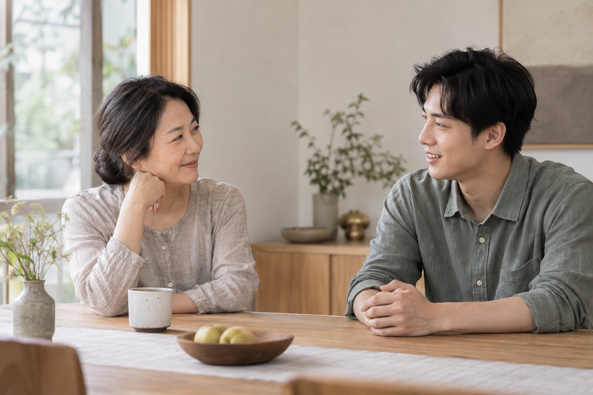Two adults in conversation across a pale wooden table beside a window.
