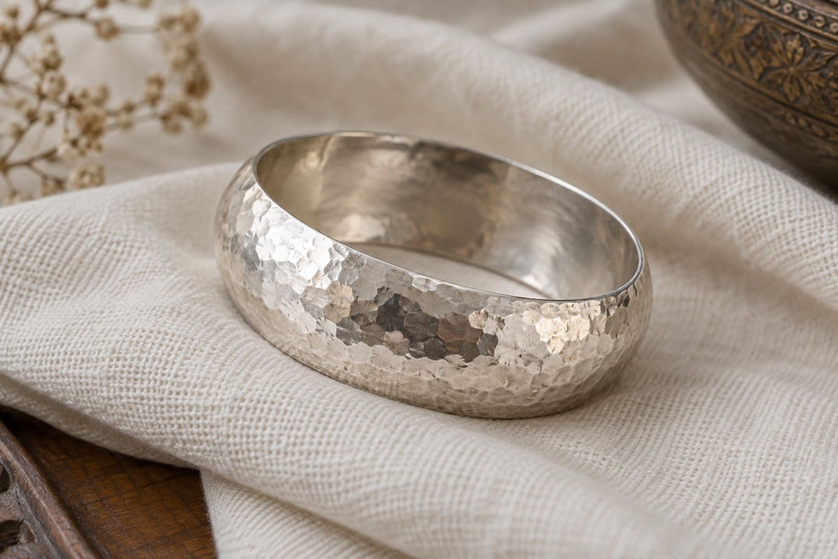 A silver bangle with small irregular hammered facets on folded cream fabric.