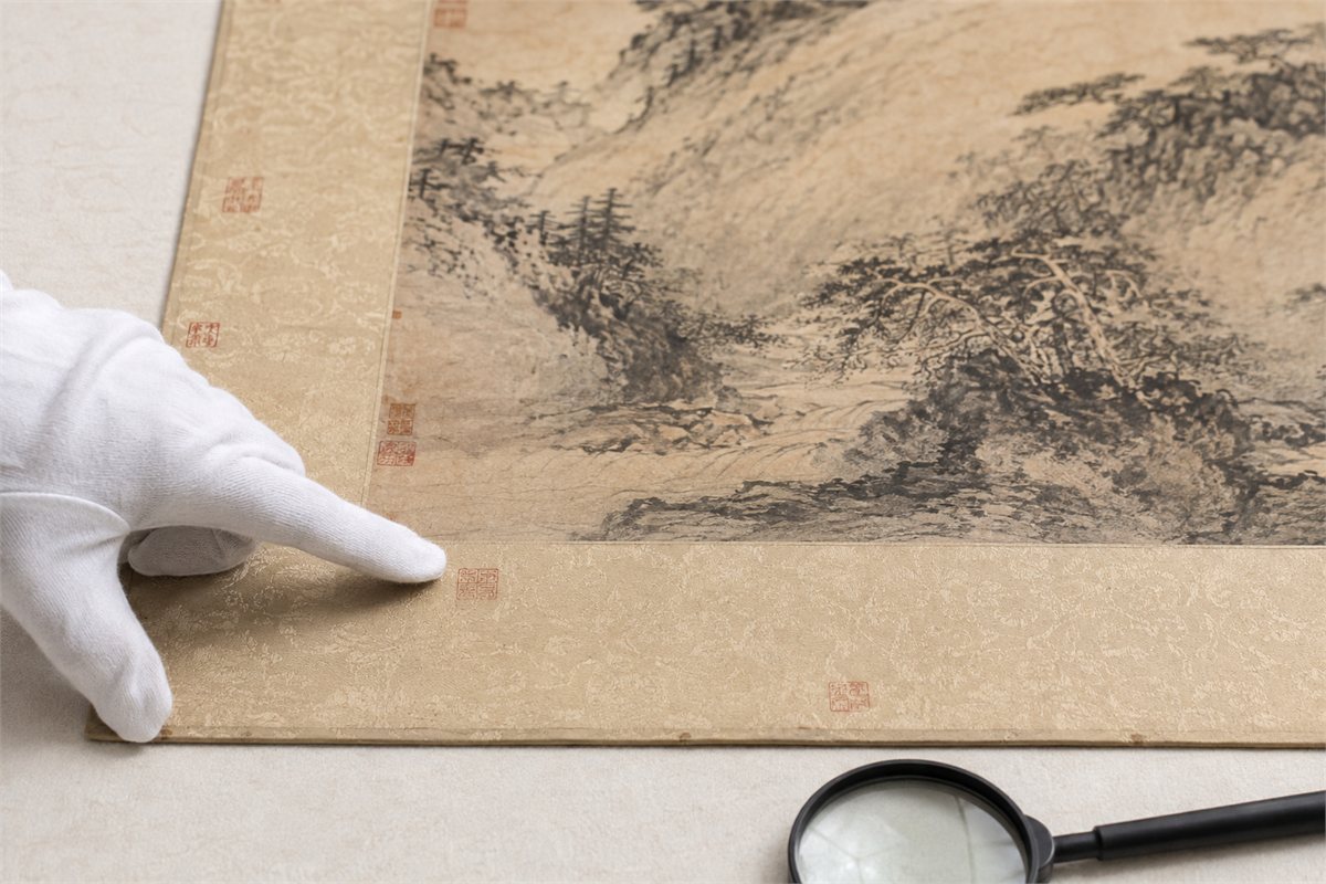 Gloved hand observing small faded seal impressions on a mounted ink painting without touching them