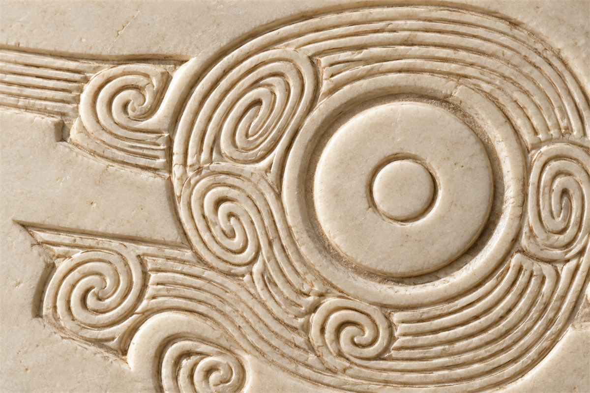 Close view of a circular eye surrounded by shallow carved curls in cream stone.