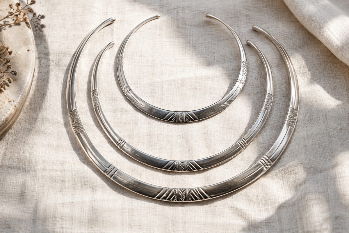 Three curved silver-colored neck ornaments arranged in overlapping layers on linen.