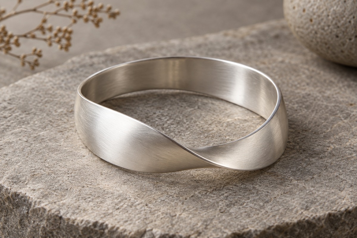 A broad silver bangle with one smooth flowing twist on a warm gray stone surface.