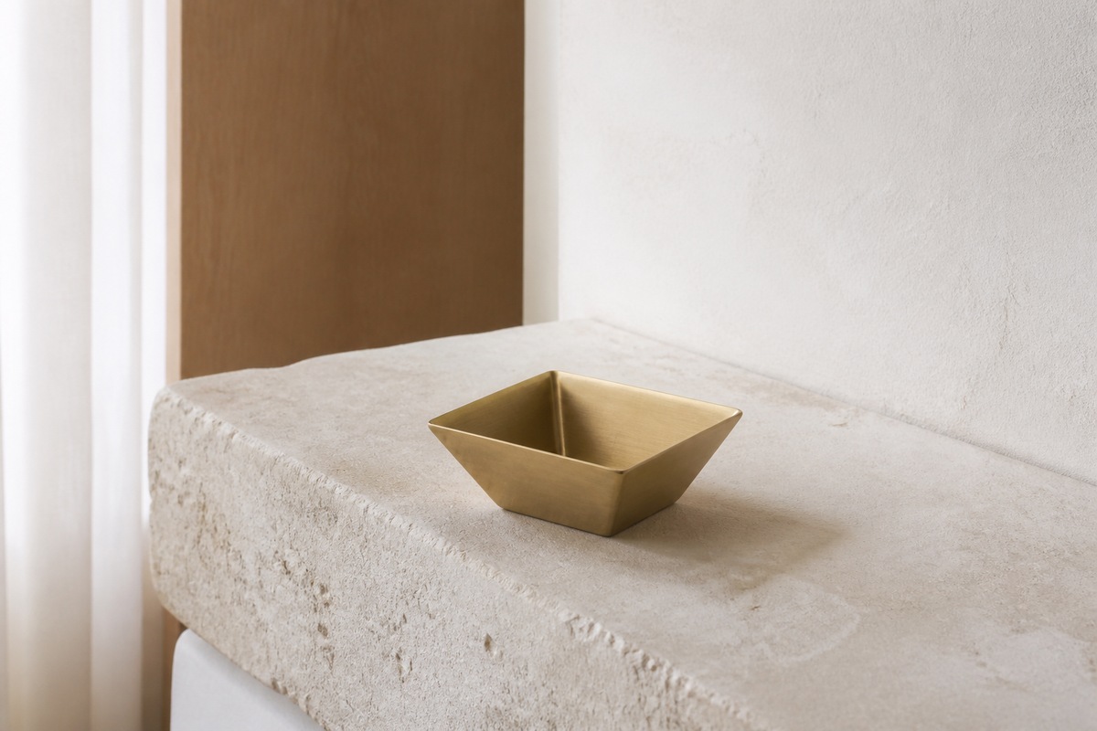 A small brass-colored square vessel stands on a pale stone shelf.