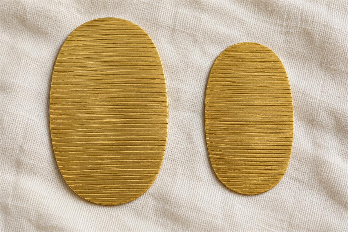 Two flat gold-colored oval shapes with fine horizontal lines on cream fabric.