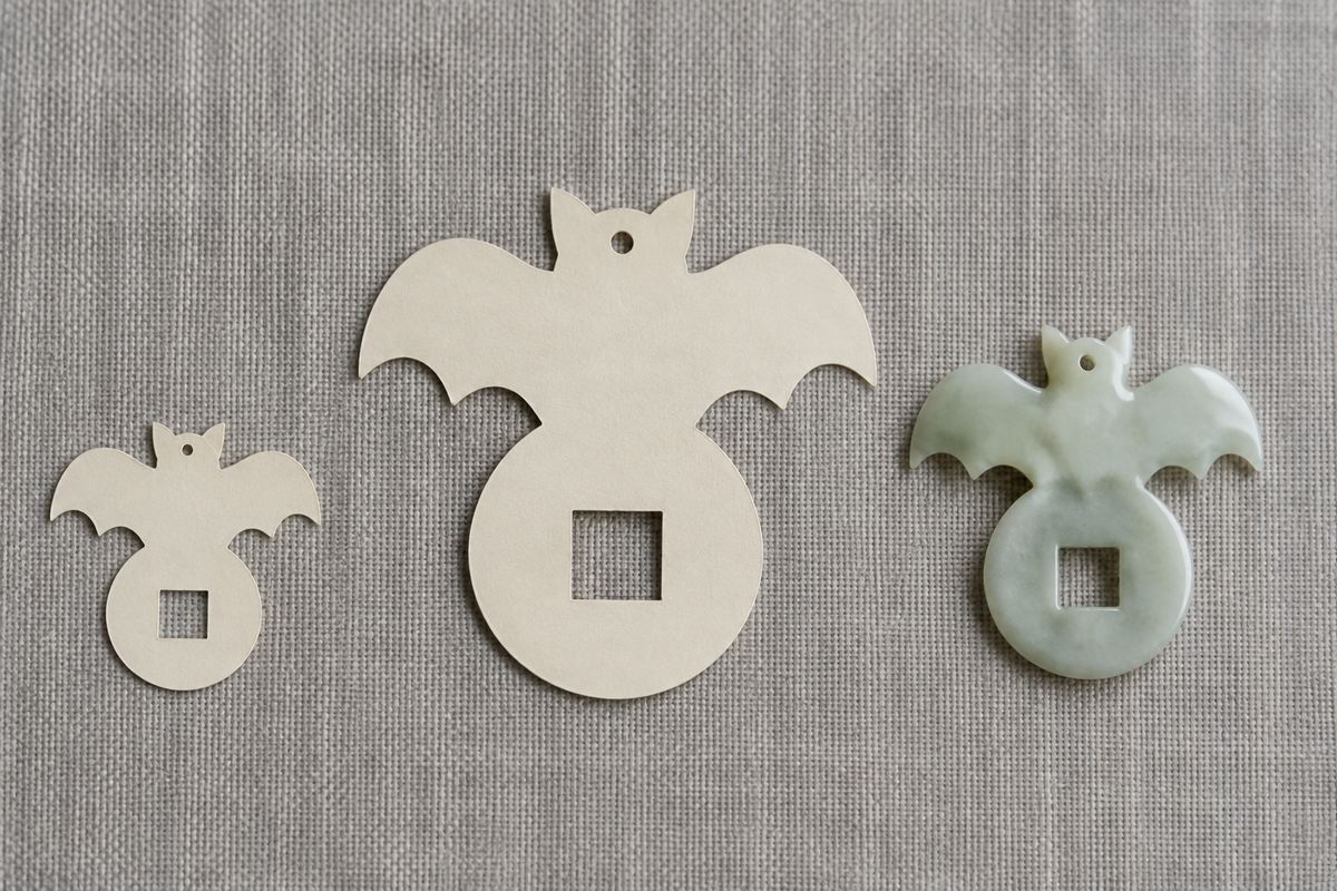 Two differently sized pendant outlines cut from plain paper beside a simple bat-and-coin pendant.