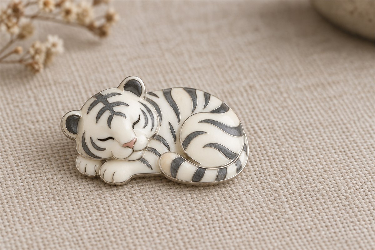 Rounded ivory and gray enamel tiger brooch resting on woven cloth.