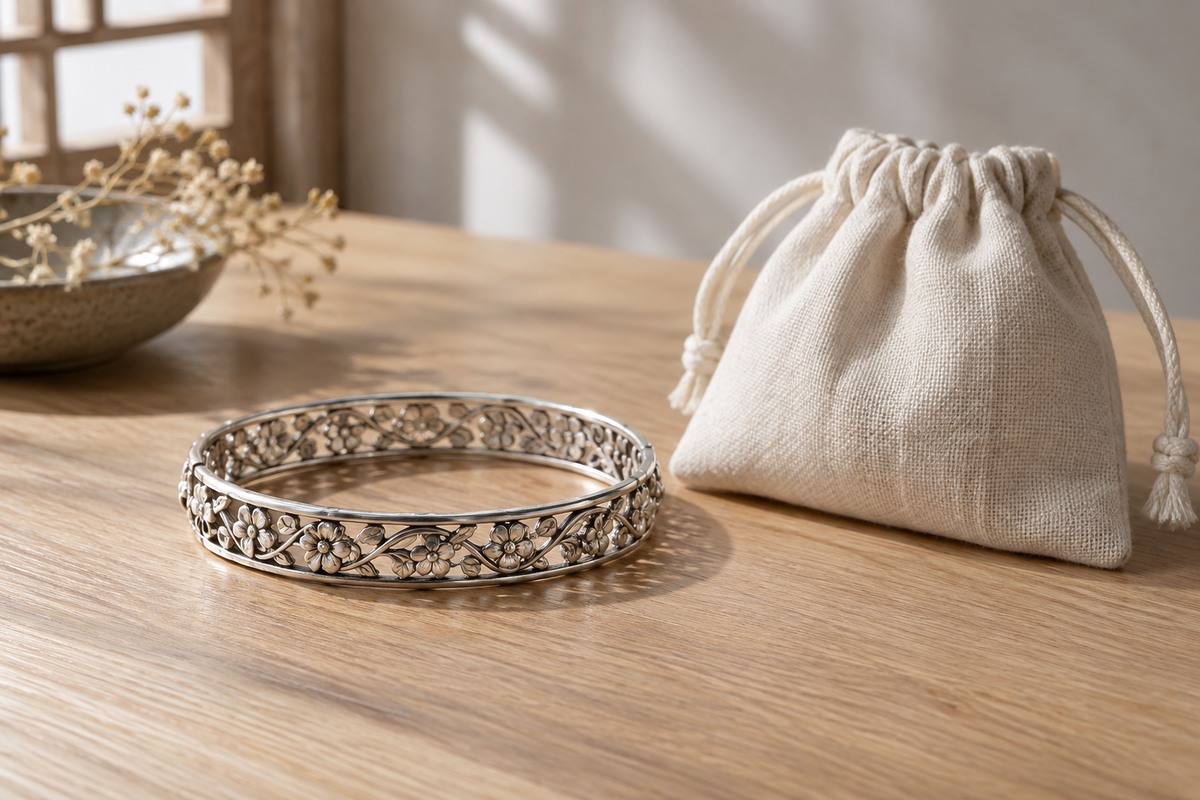 A delicate silver bangle with restrained floral openwork beside a simple cloth pouch.