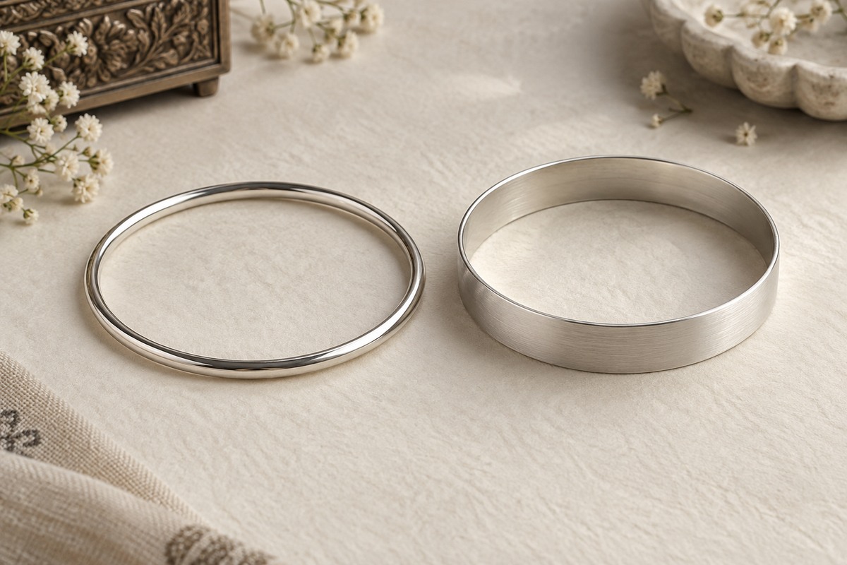 A narrow polished silver bangle and a broader satin silver bangle on warm ivory paper.