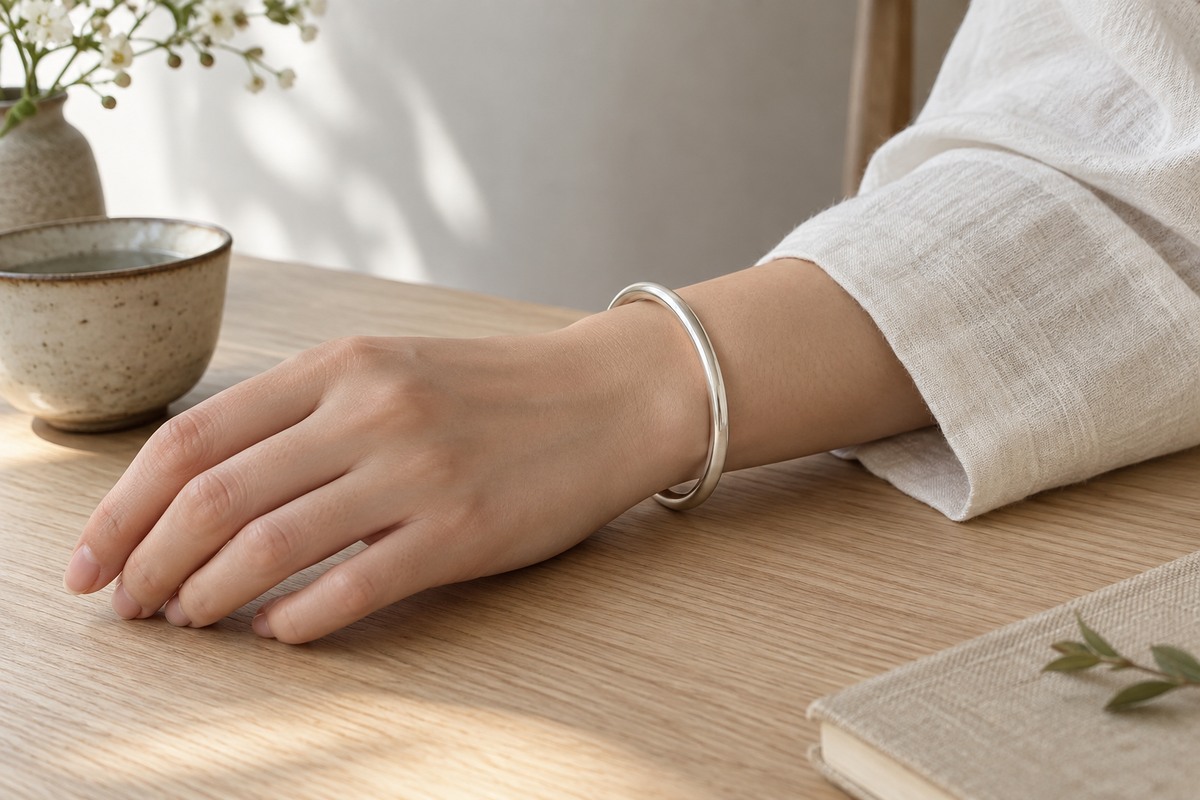 A plain silver bangle resting loosely on an adult wrist beside an ivory linen sleeve.