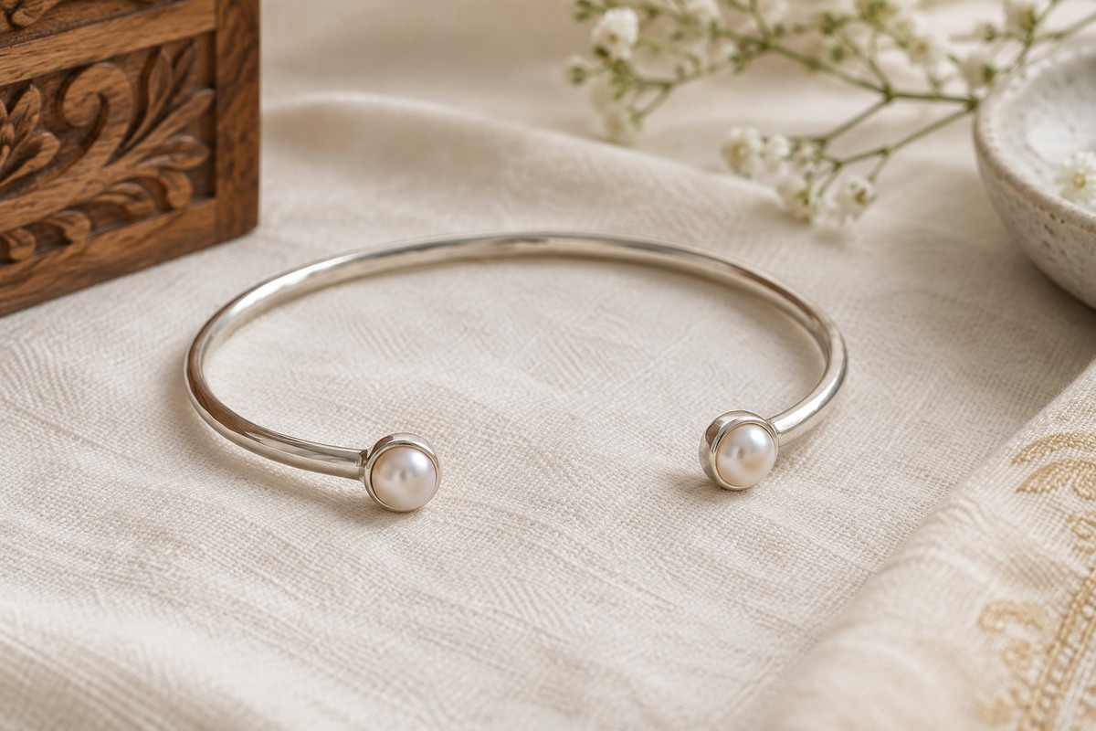 An open silver cuff with two pearl ends resting on a soft cream cloth.