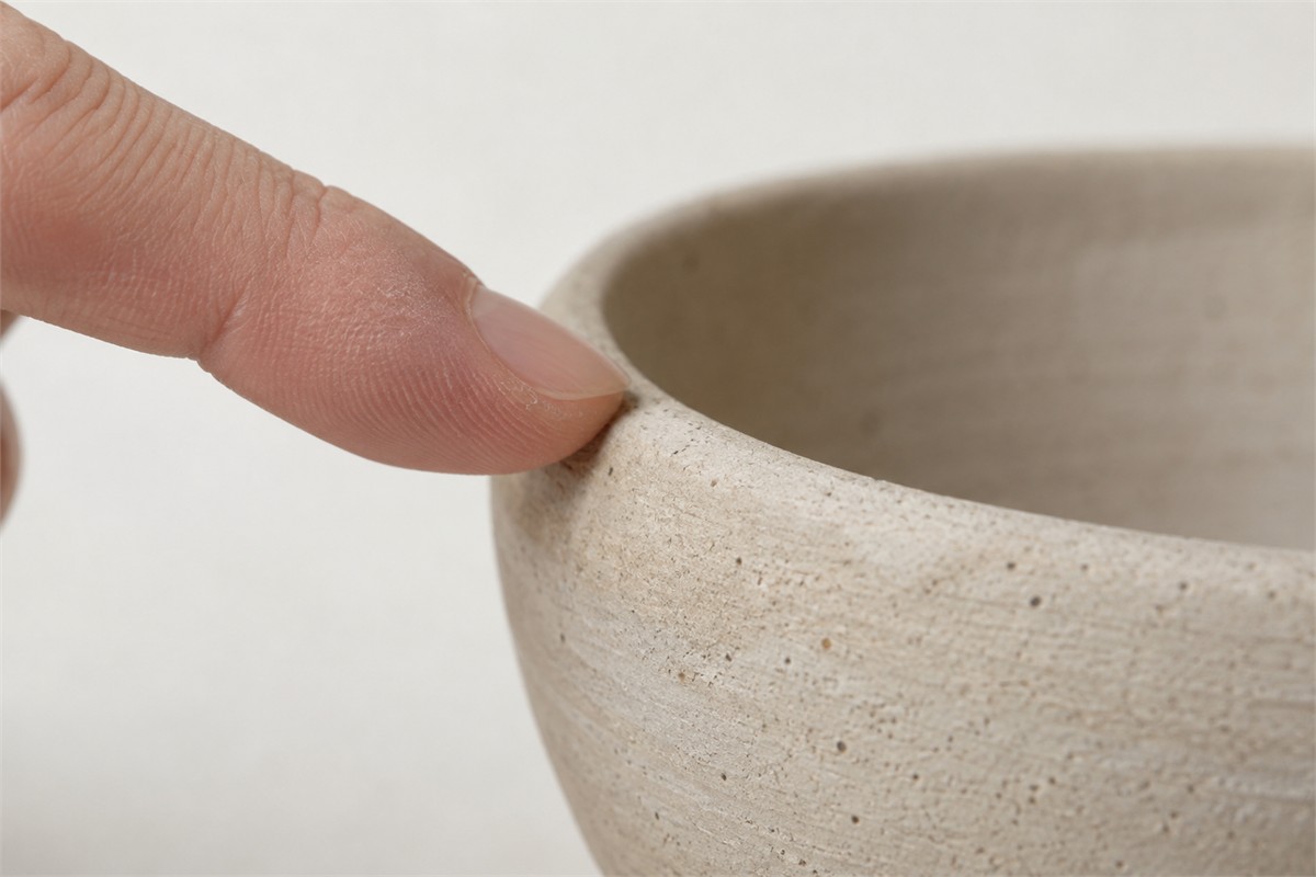 A fingertip touches the textured rim of an unglazed ceramic bowl.
