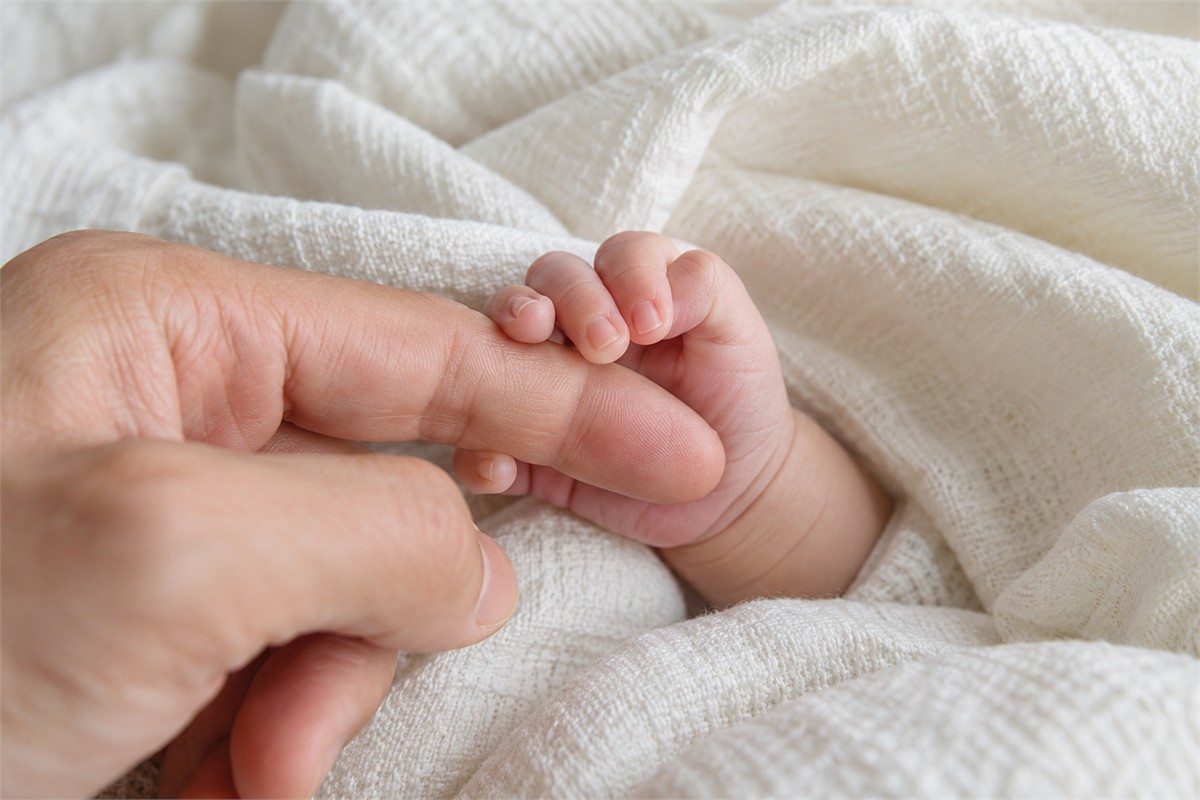 A newborn’s small hand curls around an adult’s finger over a soft blanket.